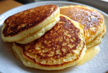 Close-up of three fluffy almond flour pancakes stacked on a white plate, drizzled with syrup.