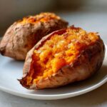Close-up of two fluffy baked sweet potatoes, one split open showing bright orange flesh and a crispy topping.