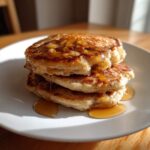 A close-up of a stack of three fluffy banana pancakes generously drizzled with golden syrup.