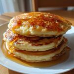 A tall stack of three fluffy banana pancakes drizzled generously with golden syrup.
