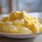 Close-up of incredibly fluffy scrambled eggs piled high on a white plate, showcasing their soft texture.