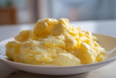 Close-up of incredibly fluffy scrambled eggs piled high on a white plate, showcasing their soft texture.