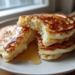 A stack of fluffy sourdough pancakes, one broken open to show the airy interior, drizzled with syrup.