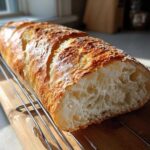 Close-up of a golden-crusted, freshly baked French bread loaf resting on a cooling rack, showing an airy interior.