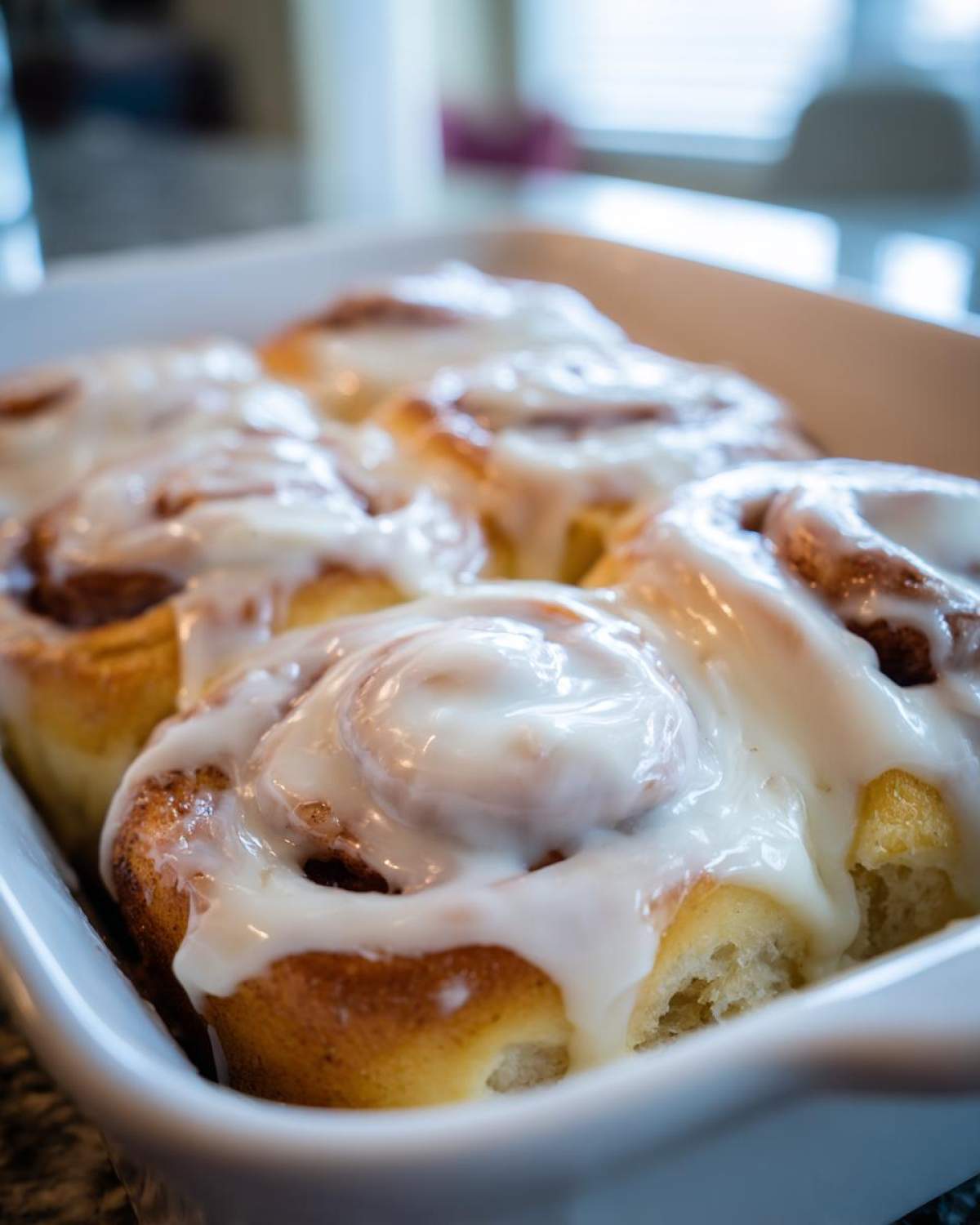Close-up of several freshly baked cinnamon roll treats covered in thick white icing in a white baking dish.
