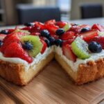 A cross-section view of a fruit pizza sugar cookie tart topped with cream cheese frosting, strawberries, kiwi, and blueberries.