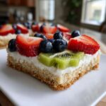 A close-up slice of fruit pizza sugar cookie topped with cream cheese frosting, strawberries, blueberries, and kiwi.