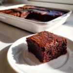 A close-up of a fudgy square of sweet potato brownies served on a white plate, with the rest of the pan visible in the background.