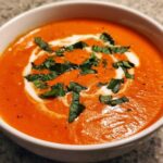 Close-up of a rich, orange-red bowl of tomato bisque, garnished with a swirl of cream and fresh basil.