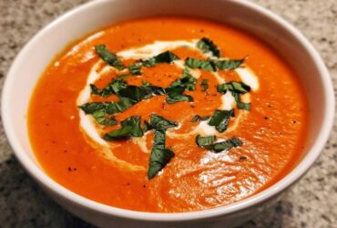Close-up of a rich, orange-red bowl of tomato bisque, garnished with a swirl of cream and fresh basil.