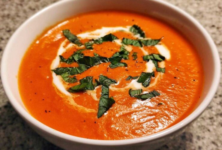 Close-up of a rich, orange-red bowl of tomato bisque, garnished with a swirl of cream and fresh basil.