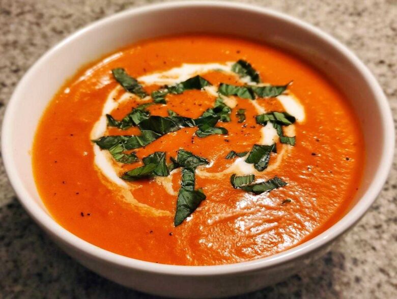 Close-up of a rich, orange-red bowl of tomato bisque, garnished with a swirl of cream and fresh basil.