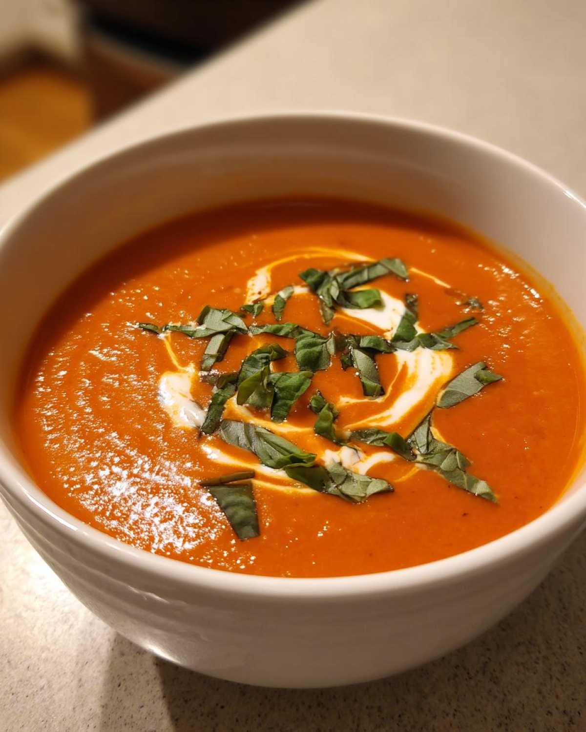 A close-up of a bowl of vibrant orange tomato bisque, swirled with cream and topped with fresh basil.