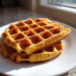 Close-up of a stack of golden brown high protein waffles resting on a white plate, backlit by sunlight.