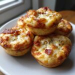 A stack of four freshly baked, golden brown pizza muffins topped with melted cheese and herbs on a white plate.