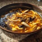 Close-up of a rich, dark broth filled with egg ribbons and mushrooms in a bowl of hot and sour soup recipe.