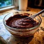 A glass bowl filled with thick, rich homemade chocolate pudding mixture being whisked.
