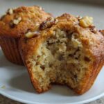 Close-up of moist banana nut muffins, one bitten open to show the texture, topped with walnuts.