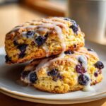Two golden-brown blueberry scones stacked on a plate, drizzled generously with white vanilla glaze.