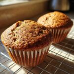 Two freshly baked, moist bran muffins cooling on a wire rack, highlighted by warm sunlight.