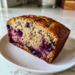 A close-up of a moist slice of blueberry banana bread served on a small white plate.