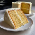 A close-up of a thick slice of moist vanilla cake recipe with creamy white frosting layers on a white plate.