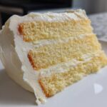 Close-up of a moist slice from a three-layer vanilla cake recipe, covered in thick white buttercream frosting.