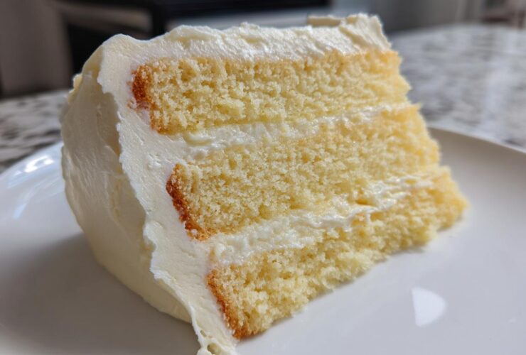 Close-up of a moist slice from a three-layer vanilla cake recipe, covered in thick white buttercream frosting.
