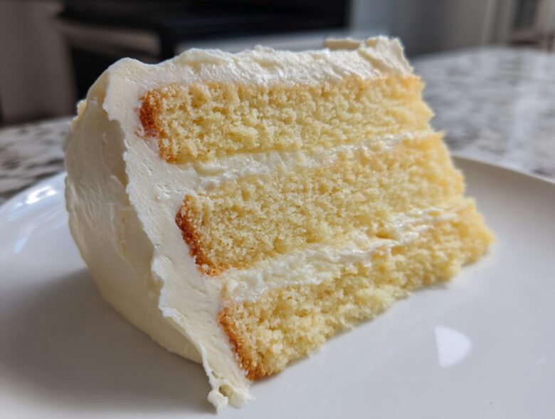 Close-up of a moist slice from a three-layer vanilla cake recipe, covered in thick white buttercream frosting.