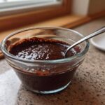 Close-up of rich, dark homemade chocolate pudding in a clear glass bowl with a spoon resting inside.