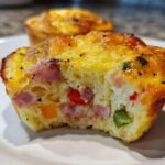 A cross-section of a savory Egg Muffin Cup showing chunks of ham, red, orange, and green peppers baked into the fluffy egg base.