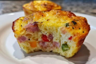 A cross-section of a savory Egg Muffin Cup showing chunks of ham, red, orange, and green peppers baked into the fluffy egg base.