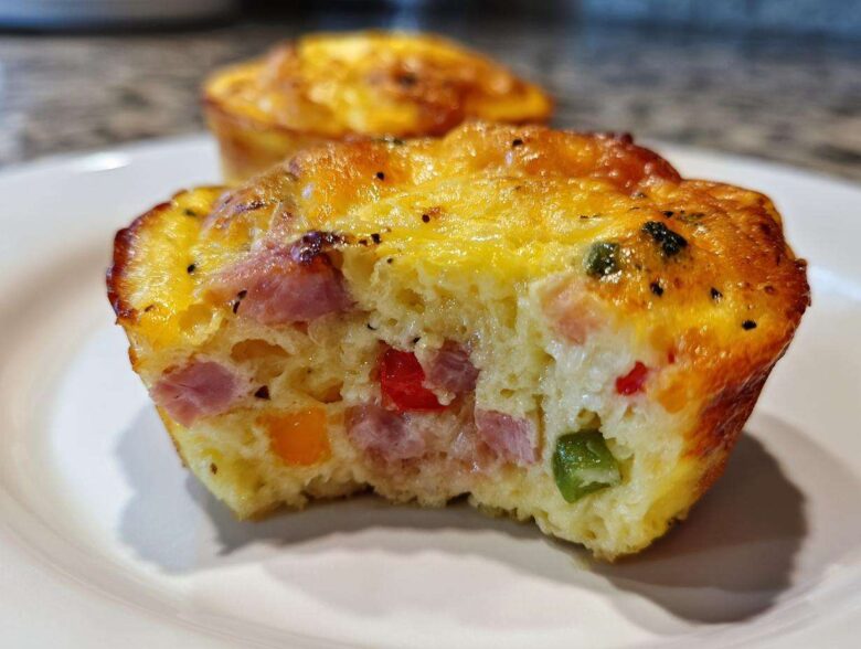 A cross-section of a savory Egg Muffin Cup showing chunks of ham, red, orange, and green peppers baked into the fluffy egg base.