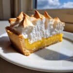 A close-up of a slice of lemon meringue tart showing thick yellow curd and toasted meringue peaks.