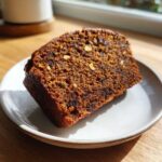 Close-up of a thick slice of moist date nut bread, showing visible chopped nuts and dark fruit pieces.
