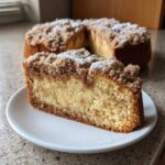A delicious slice of coffee cake with a thick cinnamon streusel topping and powdered sugar, with the rest of the coffee cake blurred in the background.
