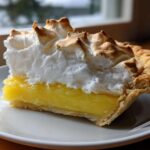 A thick slice of lemon meringue pie showing bright yellow filling and tall, torched meringue topping.
