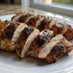 Close-up of sliced, herb-crusted greek chicken breast showing juicy white meat and a browned exterior.