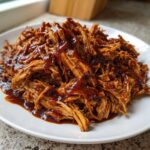 Close-up of tender, shredded slow cooker pulled pork generously coated in rich barbecue sauce on a white plate.