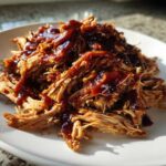 A mound of shredded, tender Slow Cooker Pulled Pork drizzled generously with dark BBQ sauce on a white plate.