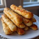 A close-up stack of golden brown, soft garlic breadsticks seasoned with herbs on a white plate.