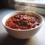 A close-up of a steaming white bowl filled with rich, thick crockpot chili featuring ground beef and kidney beans.