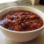 Close-up of a steaming white bowl filled with rich, hearty crockpot chili featuring ground beef and kidney beans.