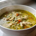A close-up of a steaming white bowl filled with rich, yellow penicillin soup, featuring shredded chicken, carrots, and fresh parsley.