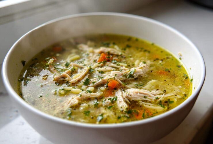 A close-up of a steaming white bowl filled with rich, yellow penicillin soup, featuring shredded chicken, carrots, and fresh parsley.