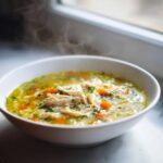 A close-up of a steaming white bowl filled with rich penicillin soup, featuring shredded chicken, rice, carrots, and parsley.