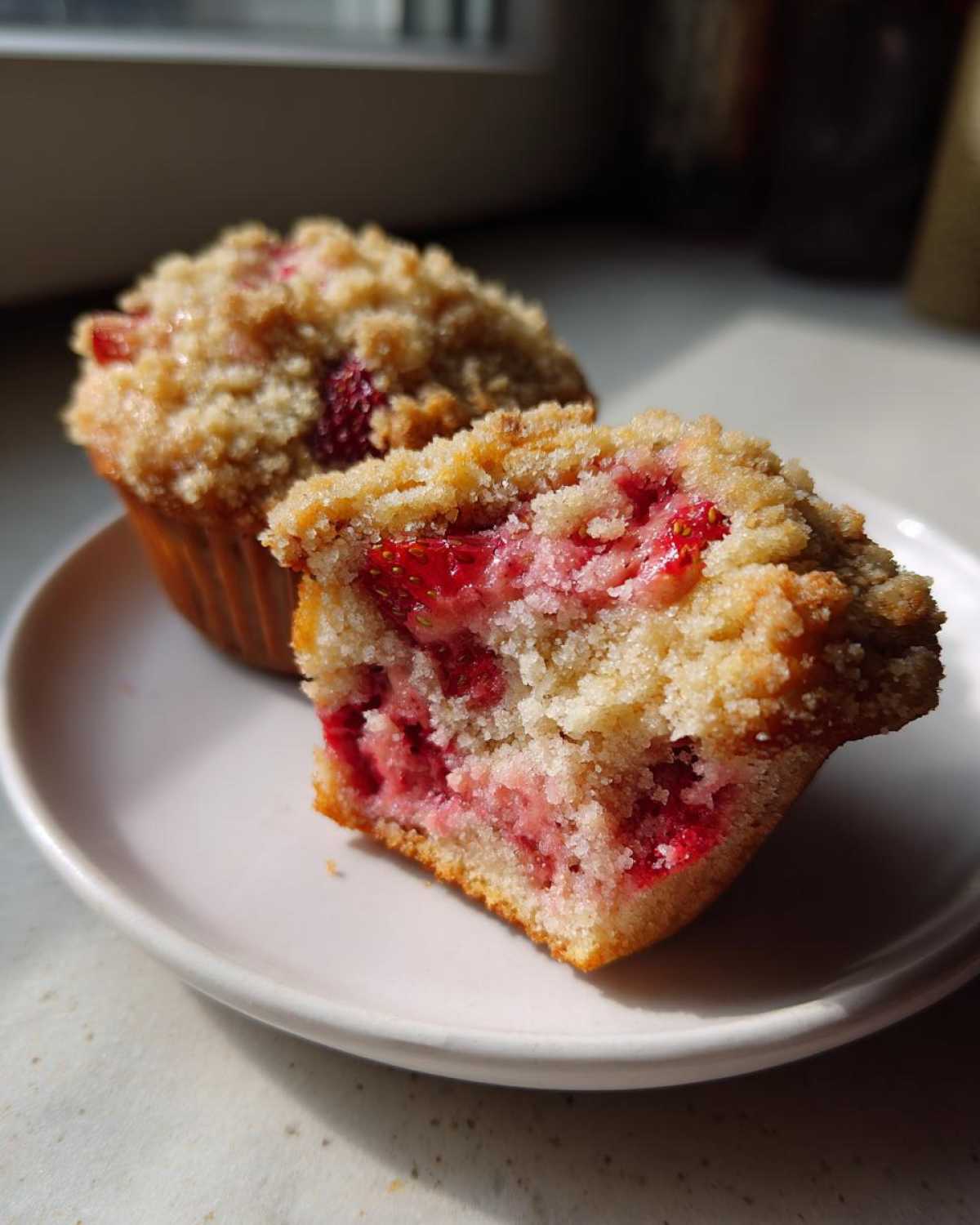 Close-up of a strawberry muffin cut in half showing moist interior and fresh strawberries, next to a whole strawberry muffin.
