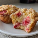 A close-up of a halved strawberry muffin showing the moist interior packed with strawberries and a sugary crumb topping.