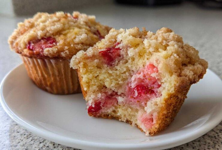 A close-up of a halved strawberry muffin showing the moist interior packed with strawberries and a sugary crumb topping.