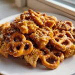 A pile of crunchy pretzels and cereal coated in shiny toffee glaze, sprinkled with coarse sugar, part of the toffee chex mix recipe.